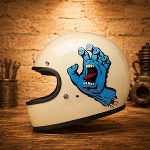 Peut inclure: Un casque de moto intégral de couleur crème arborant un motif de main bleue hurlante avec une langue rouge et l'inscription "Santa Cruz" sur le côté. Le casque présente une bordure noire le long de la base et autour de l'ouverture faciale.