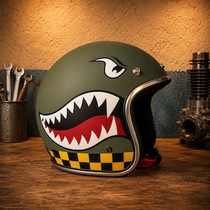May include: An olive green open-face motorcycle helmet featuring a graphic of a stylized shark mouth with sharp white teeth and a red interior. The helmet includes a black and yellow checkered pattern along the bottom edge and a silver trim.