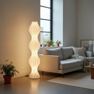 May include: A tall, cream-colored floor lamp with a unique, stacked, organic design. The lamp has a ribbed texture and emits a warm, inviting glow. The lamp is approximately 60 inches tall and is in a modern living room setting.