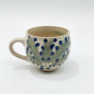 Puede incluir: Una taza de cerámica de color crema con forma redondeada. La taza presenta un diseño pintado a mano de hojas verdes y bayas azules. Tiene un asa curva y es adecuada para bebidas calientes.