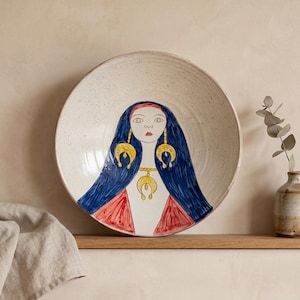 May include: A round ceramic bowl with a speckled off-white glaze featuring a hand-painted illustration of a person with long dark blue hair, red clothing, and gold-colored jewelry including large crescent-shaped earrings and a matching necklace.