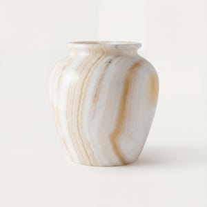 May include: A decorative vase with a rounded body and a narrow neck. The vase is made of a white and tan-colored stone, with swirling patterns throughout. The vase is a neutral color and would complement any decor.