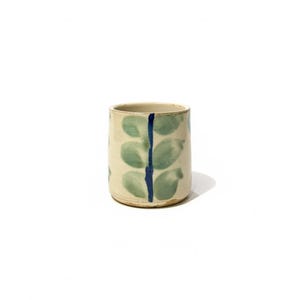 Puede incluir: Un vaso de cerámica con base color crema, adornado con un patrón de formas de hojas verdes y una franja vertical azul central. La taza hecha a mano es cilíndrica y adecuada para bebidas.