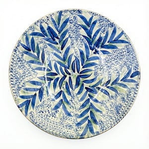 May include: A large, round ceramic plate with a hand-painted design. The plate features a central pattern of blue and green leaves against a speckled white and blue background. The rim has a gold-coloured trim.