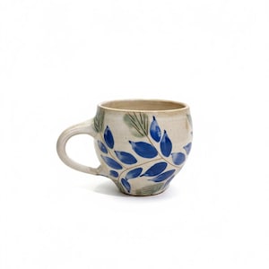 May include: A ceramic mug with a rounded shape and a handle. The mug is beige with blue leaf designs and green accents. Ideal for tea or coffee, this mug is a charming addition to any kitchen.