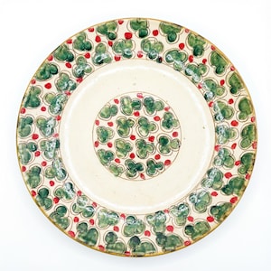 May include: A circular ceramic plate with a cream-coloured centre and a decorative border. The border features a pattern of green clover-like shapes and small red dots. The rim has a gold-coloured trim. The plate is likely for serving food.