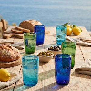 May include: A collection of six cylindrical drinking glasses in varying shades of blue and green, each featuring a textured, bubbly surface. The glasses are arranged on a wooden table alongside loaves of bread, bowls of olives, and lemons.