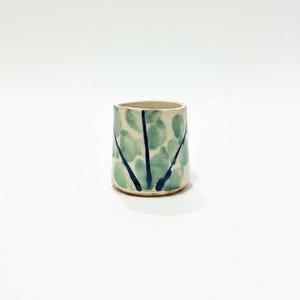 May include: A ceramic tumbler with a cream base, decorated with light green, leaf-like shapes and dark blue vertical lines. The handmade cup is cylindrical with a slightly flared rim.