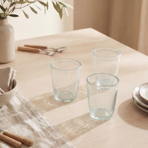 May include: Three clear glass tumblers with a subtle bubbled texture. The glasses are cylindrical with a slightly tapered base, featuring a light, translucent appearance that catches the natural light on the surface.