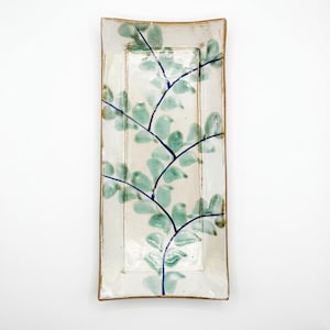 Peut inclure: Plat à servir rectangulaire en céramique avec une base crème et un motif botanique peint à la main. Le motif présente des feuilles vert clair et des tiges bleu foncé. Le plat a un bord légèrement surélevé avec une bordure dorée.