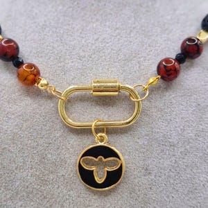 May include: A beaded necklace featuring a gold-tone clasp and a black and gold bee pendant. The necklace incorporates various beads in shades of red, black, orange, and pink. The clasp is a large, oval-shaped design.