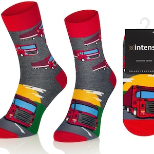Puede incluir: Calcetines grises con puños y punteras rojas, estampados con camiones rojos. Los calcetines tienen un diseño colorido en la parte inferior con una carretera, cielo amarillo y hierba verde. El embalaje muestra un camión rojo.