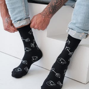 Può includere: Calzini neri con un motivo bianco di attrezzature per il sollevamento pesi. I calzini, indossati con jeans blu, presentano un motivo ripetuto di bilancieri, manubri e dischi. I calzini sono al ginocchio.