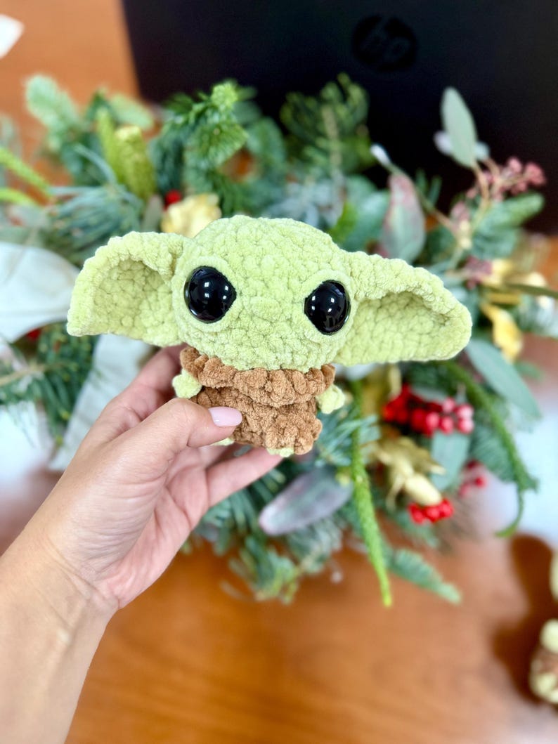 May include: A light green crocheted doll with large black eyes and oversized ears. The doll has a brown textured body and is approximately 4 inches tall. The doll is a character from a popular science fiction franchise.