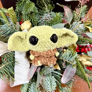 May include: A plush, light green character with large black eyes and oversized ears, nestled in a wreath of evergreen branches, red berries, and gold accents. The character has a brown outfit and is a popular character from a science fiction series.