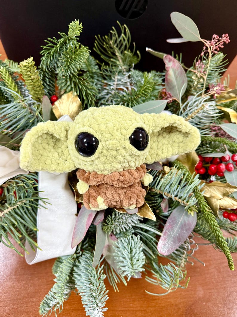 May include: A plush, light green character with large black eyes and oversized ears, nestled in a wreath of evergreen branches, red berries, and gold accents. The character has a brown outfit and is a popular character from a science fiction series.