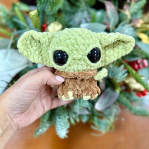 May include: A crocheted plush toy in the likeness of a character with large black eyes and oversized ears. The toy is primarily light green with a brown outfit. The toy is being held in front of a background of greenery.