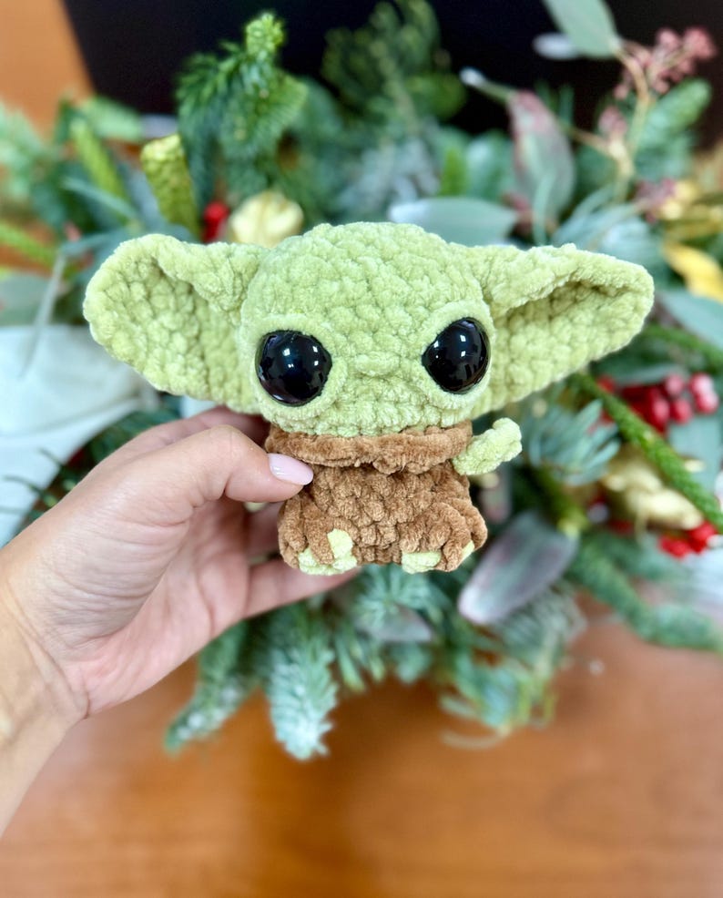 May include: A crocheted plush toy in the likeness of a character with large black eyes and oversized ears. The toy is primarily light green with a brown outfit. The toy is being held in front of a background of greenery.