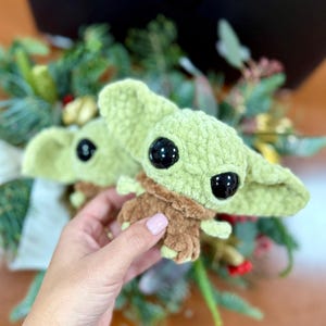 May include: Two crocheted plush figures of a character with large black eyes, green skin, and brown clothing. The figures are held in front of a blurred background of greenery and a dark surface.