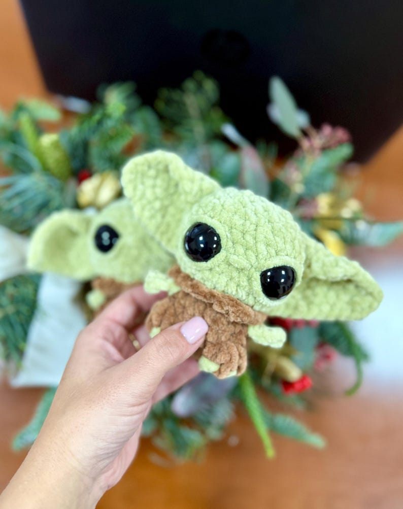 May include: Two crocheted plush figures of a character with large black eyes, green skin, and brown clothing. The figures are held in front of a blurred background of greenery and a dark surface.
