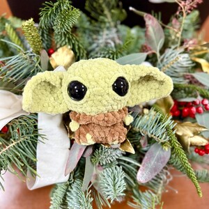 May include: A crocheted green and brown plush toy with large black eyes, resembling a character from a popular science fiction series. The toy is surrounded by a festive arrangement of evergreen branches, red berries, and decorative ribbon.