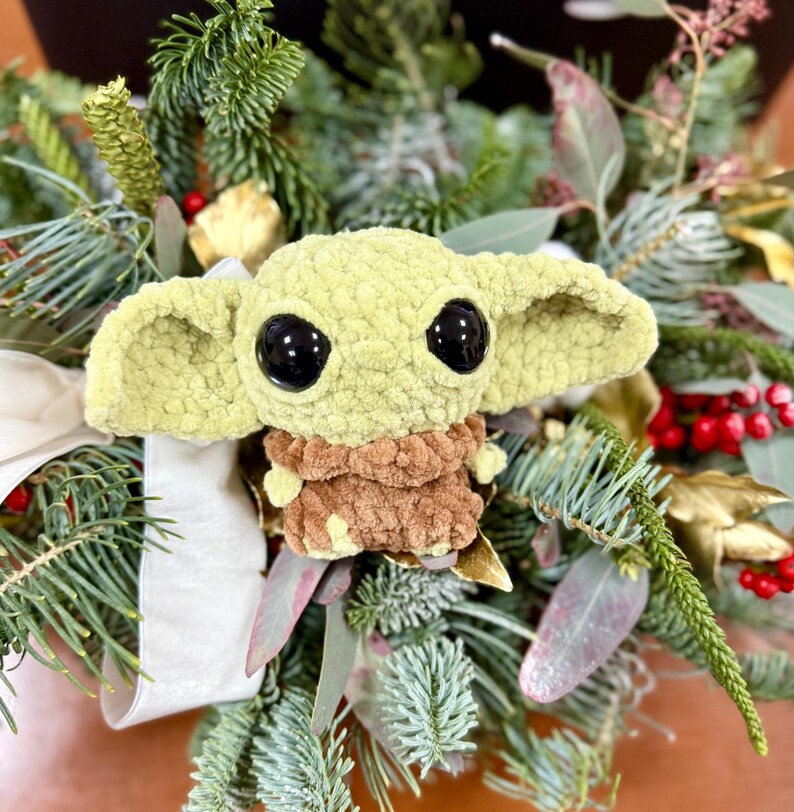 May include: A crocheted green and brown plush toy with large black eyes, resembling a character from a popular science fiction series. The toy is surrounded by a festive arrangement of evergreen branches, red berries, and decorative ribbon.