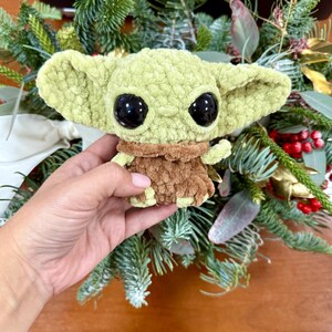 May include: A light green crocheted doll with large black eyes and a brown outfit. The doll is held in front of a background of greenery and red berries. The doll's large ears and facial features are prominent.