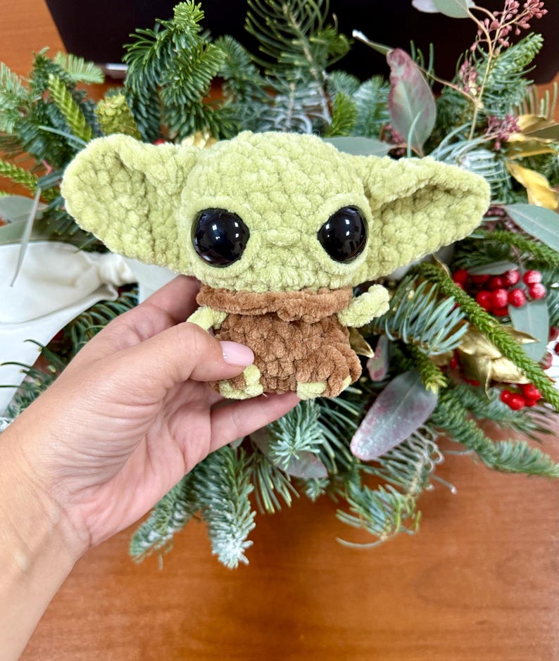 May include: A light green crocheted doll with large black eyes and a brown outfit. The doll is held in front of a background of greenery and red berries. The doll's large ears and facial features are prominent.
