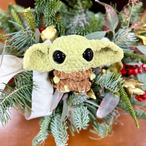 May include: A handmade, light green crocheted character with large black eyes and a brown outfit, nestled in a festive arrangement of evergreen branches, red berries, and white ribbon. The character is a popular figure from a science fiction franchise.