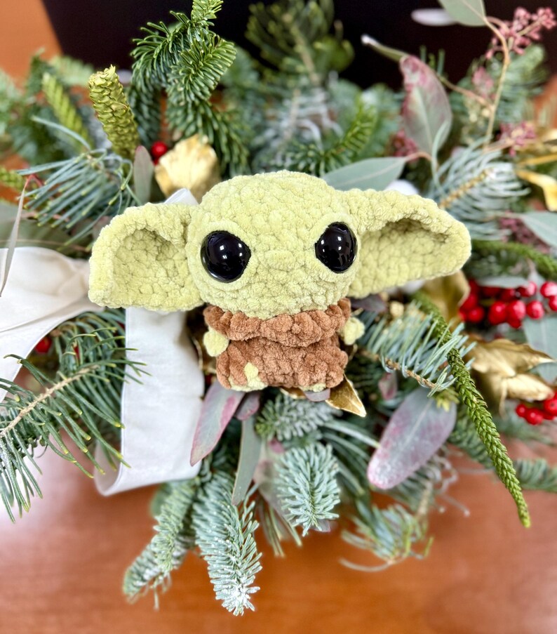 May include: A handmade, light green crocheted character with large black eyes and a brown outfit, nestled in a festive arrangement of evergreen branches, red berries, and white ribbon. The character is a popular figure from a science fiction franchise.