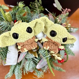 May include: Two crocheted figures of a character with large black eyes and large ears, in a light green color, with brown clothing. The figures are surrounded by a festive arrangement of evergreen branches, red berries, and gold accents.