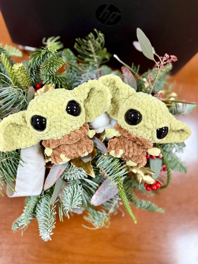 May include: Two crocheted figures of a character with large black eyes and large ears, in a light green color, with brown clothing. The figures are surrounded by a festive arrangement of evergreen branches, red berries, and gold accents.
