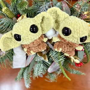 May include: Two plush green and brown character figures with large black eyes are nestled among evergreen branches, red berries, and gold accents. The figures have large ears and are surrounded by a festive holiday arrangement.