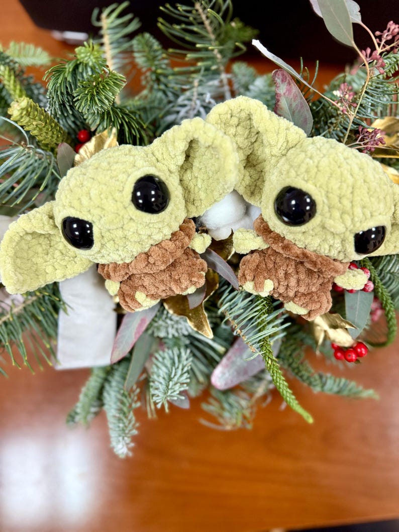 May include: Two plush green and brown character figures with large black eyes are nestled among evergreen branches, red berries, and gold accents. The figures have large ears and are surrounded by a festive holiday arrangement.