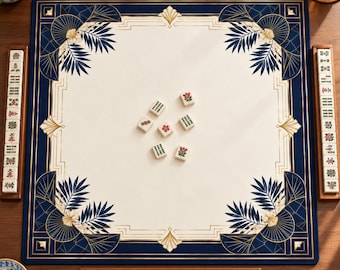 Puede incluir: Un tapete de mahjong cuadrado con un centro color crema y esquinas azul oscuro decoradas con patrones dorados de abanicos y hojas de palma. Cuatro soportes de madera sostienen fichas de mahjong, con varias fichas sueltas en el centro.