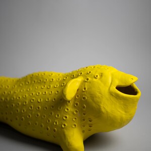 Może przedstawiać: Jasnożółta rzeźba ceramiczna stylizowanego zwierzęcia z uśmiechniętą otwartą paszczą, małymi uszami i teksturowaną powierzchnią z małymi otworami. Rzeźba na szarym tle.