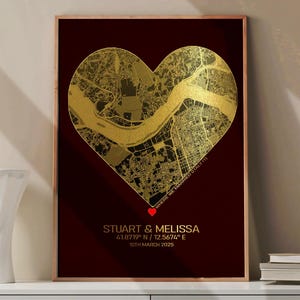 Könnte beinhalten: Gerahmter Druck mit einer goldfarbenen, herzförmigen Karte auf dunkelrotem Hintergrund. Die Karte zeigt eine Stadt und einen Fluss. Der Druck enthält die Namen "STUART & MELISSA", Koordinaten und das Datum 10. März 2025.