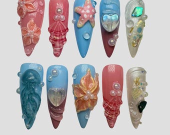 May include: A set of ten long, pointed artificial nails. Each nail features a unique design with ocean-themed embellishments, including starfish, seashells, and floral accents. Colors include pink, blue, and clear, with pearl and gem details.