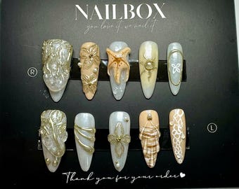 Puede incluir: Un conjunto de diez uñas artificiales con varios diseños. Las uñas presentan adornos como detalles dorados, perlas y una estrella de mar. El texto "NAILBOX you love it we nail it" y "Thank you for your order" son visibles.