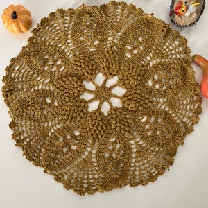 Può includere: Un centrino all'uncinetto marrone dorato fatto a mano con un intricato design floreale. Il centrino circolare presenta un motivo centrale a stella e dettagli traforati, che mostrano la delicata maestria artigianale. Oggetti decorativi circondano il centrino, tra cui una zucca, un fungo e una statuina di riccio.