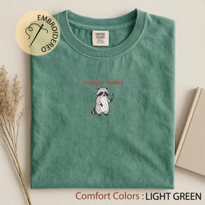 K&ouml;nnte beinhalten: Hellgr&uuml;nes T-Shirt mit einem gestickten Waschb&auml;ren, der ein Messer h&auml;lt, und dem roten Text "I require snacks". Das Shirt ist mit "Comfort Colors" gekennzeichnet und hat eine hellgr&uuml;ne Farbe.
