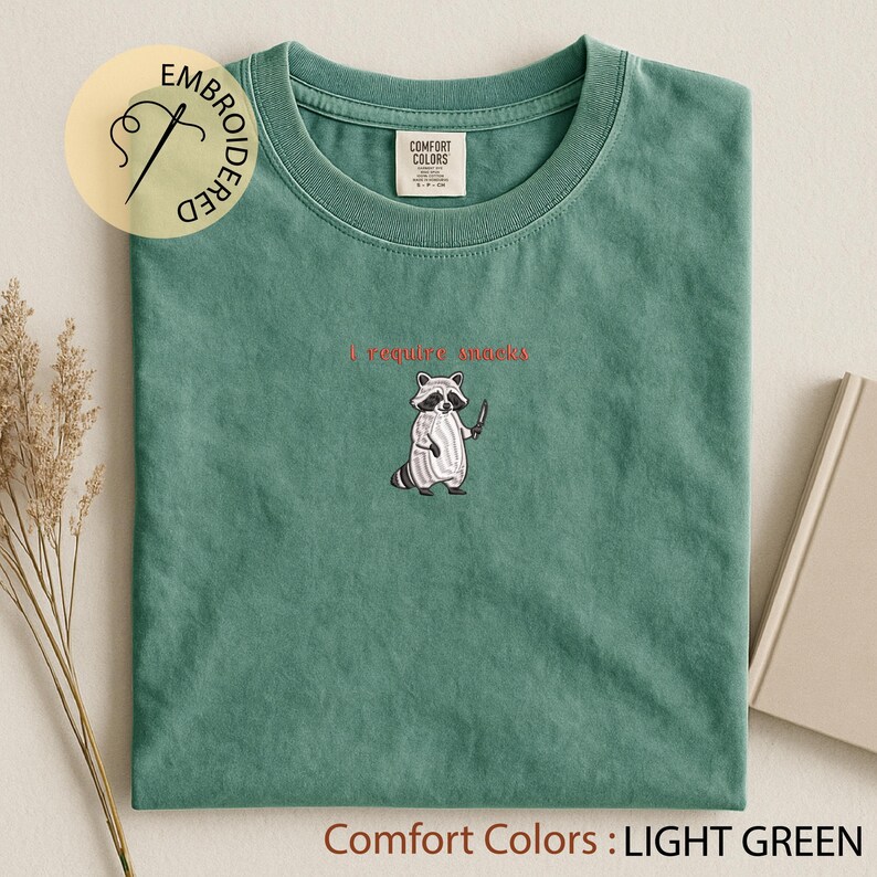 K&ouml;nnte beinhalten: Hellgr&uuml;nes T-Shirt mit einem gestickten Waschb&auml;ren, der ein Messer h&auml;lt, und dem roten Text "I require snacks". Das Shirt ist mit "Comfort Colors" gekennzeichnet und hat eine hellgr&uuml;ne Farbe.