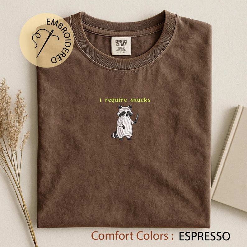 K&ouml;nnte beinhalten: Espressofarbenes T-Shirt mit Rundhalsausschnitt. Das Shirt zeigt einen gestickten Waschb&auml;ren mit einem Messer und dem Text "I require snacks" dar&uuml;ber. Das Shirt ist mit "Comfort Colors" gekennzeichnet.