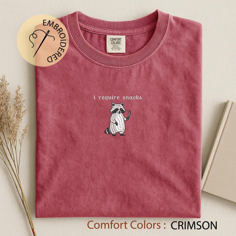 K&ouml;nnte beinhalten: Karmesinrotes T-Shirt mit Rundhalsausschnitt. Das Shirt zeigt einen gestickten Waschb&auml;ren, der ein Messer h&auml;lt, mit dem Text "i require snacks" dar&uuml;ber. Das Shirt ist mit "Comfort Colors" gekennzeichnet.