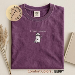 K&ouml;nnte beinhalten: Beerenfarbenes T-Shirt mit Rundhalsausschnitt. Das Shirt zeigt einen gestickten Waschb&auml;ren mit einem Messer und dem Schriftzug "i require snacks" dar&uuml;ber. Das Shirt ist mit "Comfort Colors" gekennzeichnet.