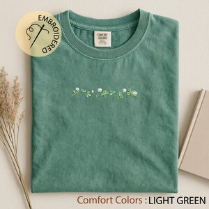 Pu&ograve; includere: T-shirt verde chiaro con ricamo floreale bianco e verde. La maglietta ha un girocollo e maniche corte. Il testo "Comfort Colors: LIGHT GREEN" &egrave; nella parte inferiore dell'immagine. Un logo circolare con la scritta "EMBROIDERED" si trova in alto a sinistra.