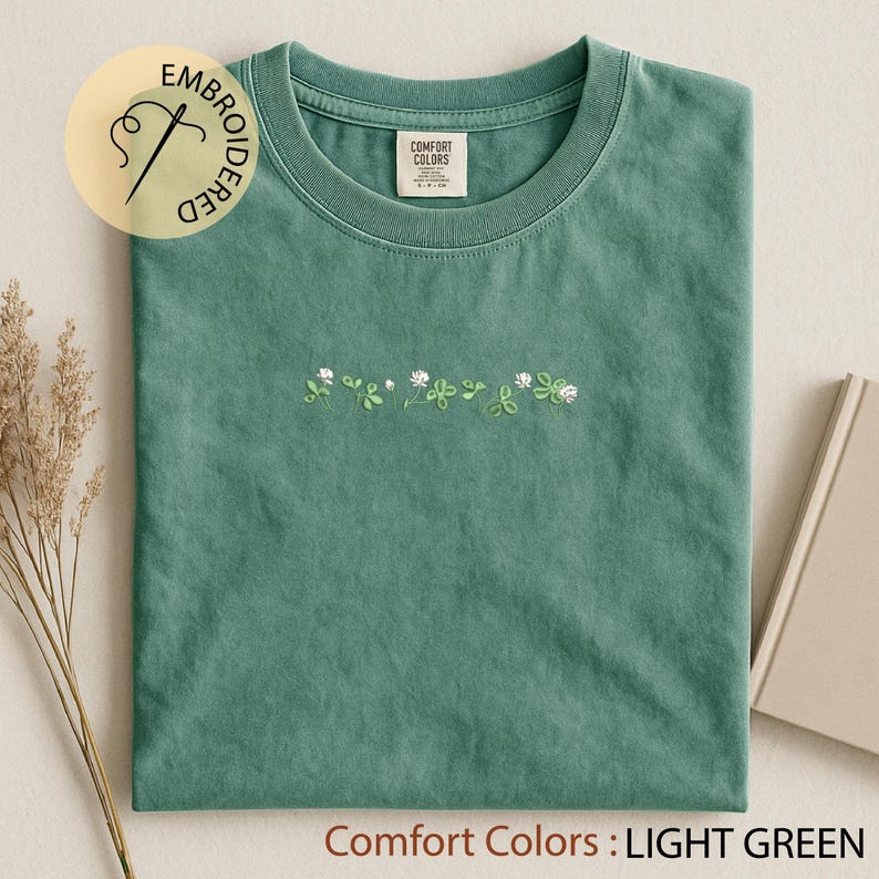 Pu&ograve; includere: T-shirt verde chiaro con ricamo floreale bianco e verde. La maglietta ha un girocollo e maniche corte. Il testo "Comfort Colors: LIGHT GREEN" &egrave; nella parte inferiore dell'immagine. Un logo circolare con la scritta "EMBROIDERED" si trova in alto a sinistra.