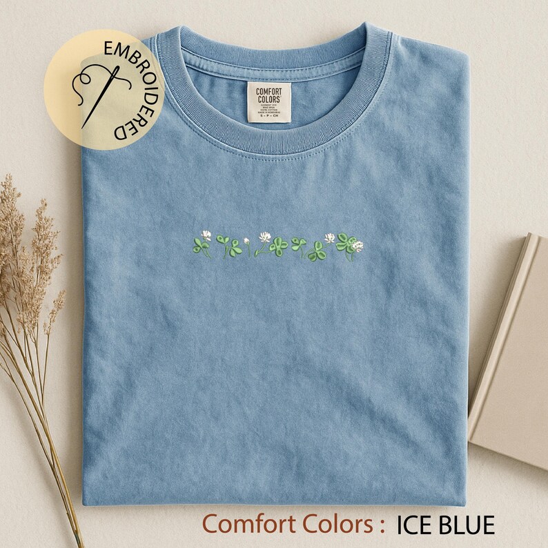 Pu&ograve; includere: T-shirt Comfort Colors blu ghiaccio con ricami di trifogli verdi e fiori bianchi. La maglietta ha un girocollo e una piccola etichetta sul colletto. Il testo "Comfort Colors: ICE BLUE" &egrave; in basso.