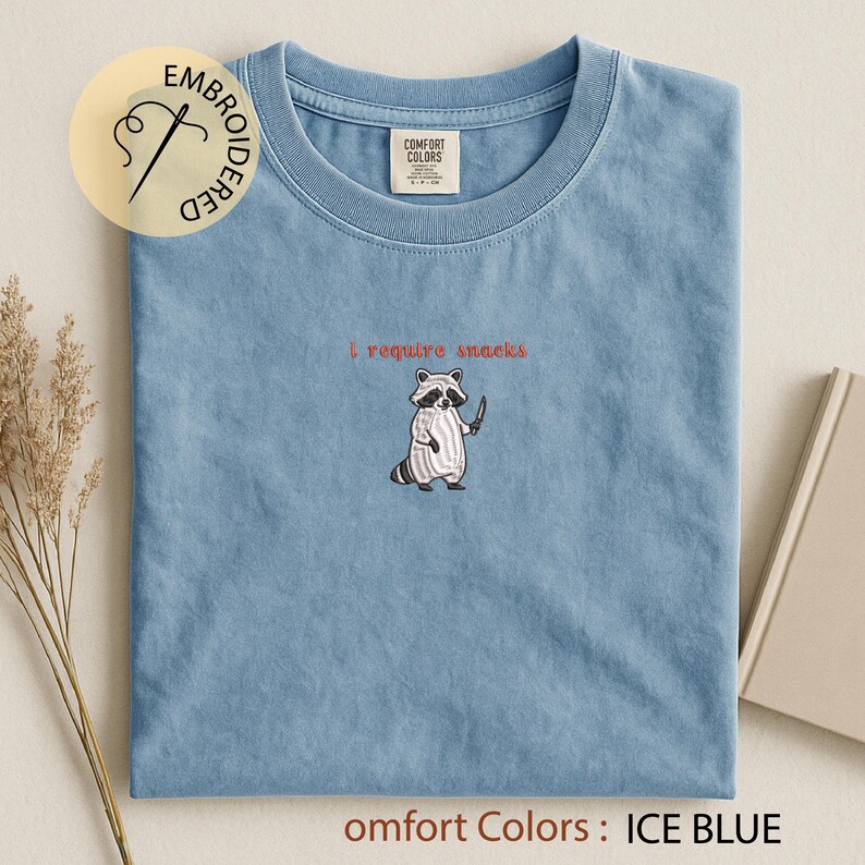 K&ouml;nnte beinhalten: Eisblaues T-Shirt mit einem Waschb&auml;r-Motiv, das ein Messer h&auml;lt, und dem roten Text "I require snacks". Das Shirt hat ein Comfort Colors-Etikett und den Text "Comfort Colors: ICE BLUE".