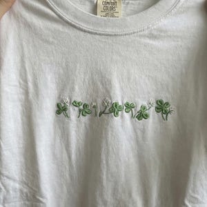 Pu&ograve; includere: T-shirt bianco con trifogli verdi ricamati e fiori bianchi sul petto. La maglietta &egrave; realizzata in un materiale morbido e confortevole. L'etichetta recita "Comfort Colors".